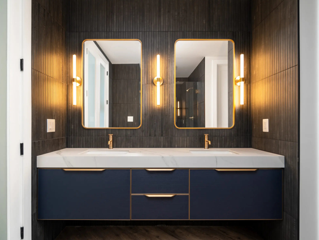 Dark and moody primary bathroom remodel featuring a double vanity with sleek cabinetry, matte black fixtures, large mirrors, and soft ambient lighting creating a modern, high-end atmosphere.